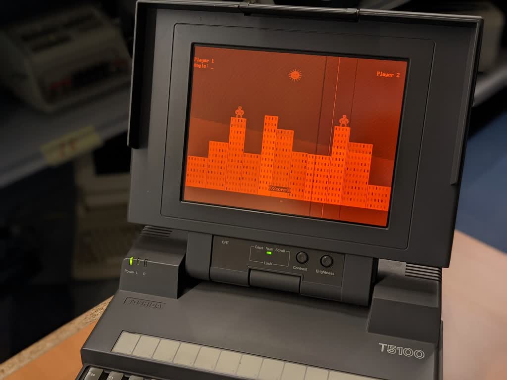 Toshiba T5100 laptop with an amber orange display running QBasic GORILLAS.BAS- monkeys standing on buildings that throw bananas to destroy each other